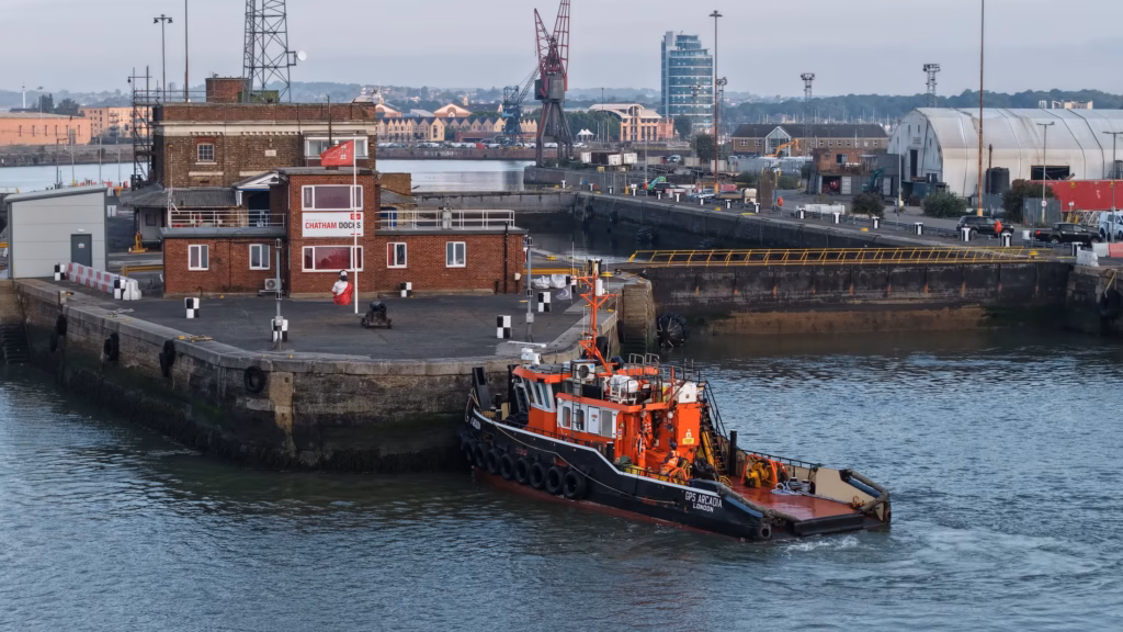 Aerial view of the tug GPS Arcadia pushing on to the dockyard wall at Chatham, with the historic dockyard buildings and cranes behind