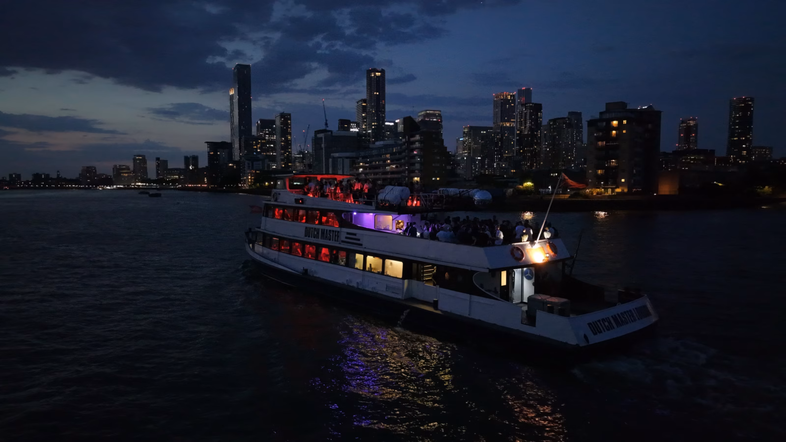 The Dutch Master under way after dark on the River Thames, lit interior decks reflecting on the water with the Canary Wharf skyline behind
