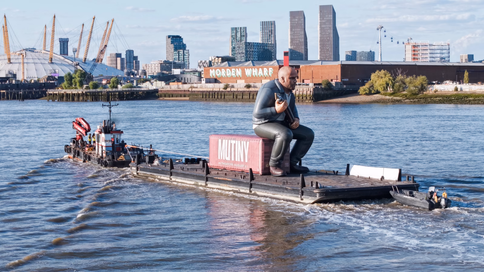 Mutiny: Jason Statham Brand Activation on the Thames 1 The multicat Emilia D towing a pontoon carrying a giant inflatable Jason Statham seated on a pink Mutiny shipping container past the Thames foreshore in London