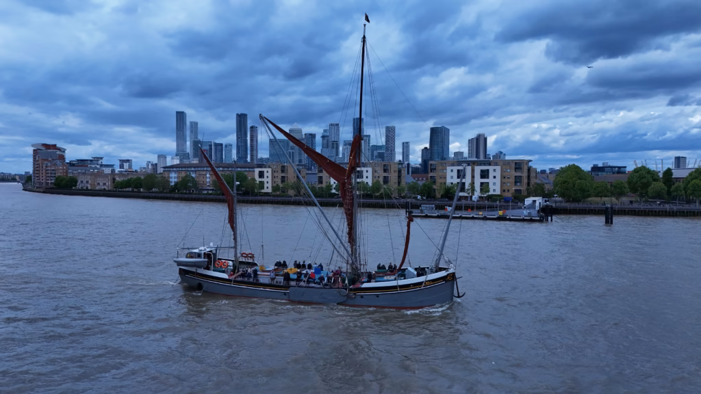 The Thames sailing barge Will under way on the tideway with passengers on deck and the Canary Wharf towers behind