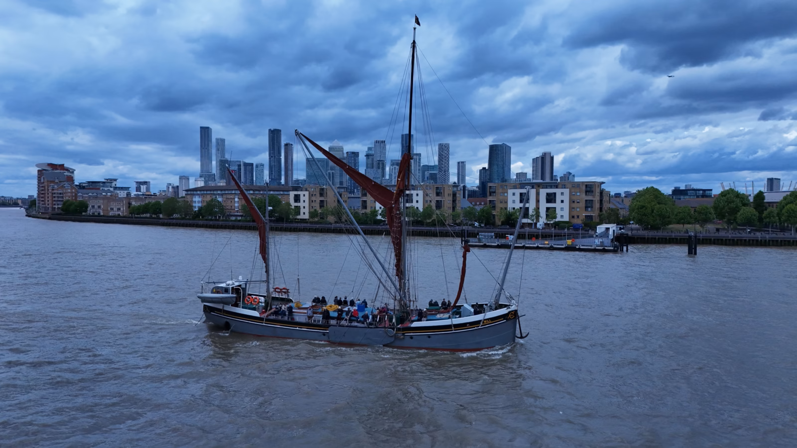 The Thames sailing barge Will under way on the tideway with passengers on deck and the Canary Wharf towers behind