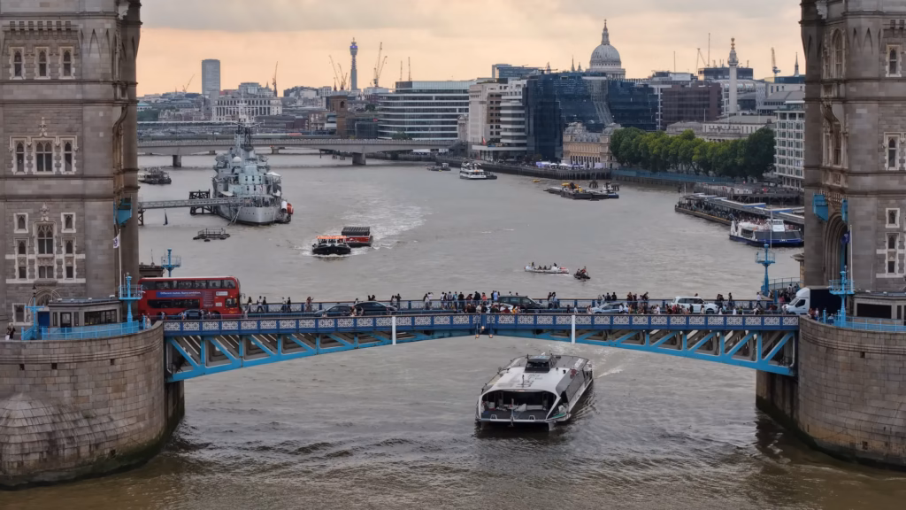A red London bus crossing Tower Bridge with HMS Belfast and the City skyline beyond, aerial view