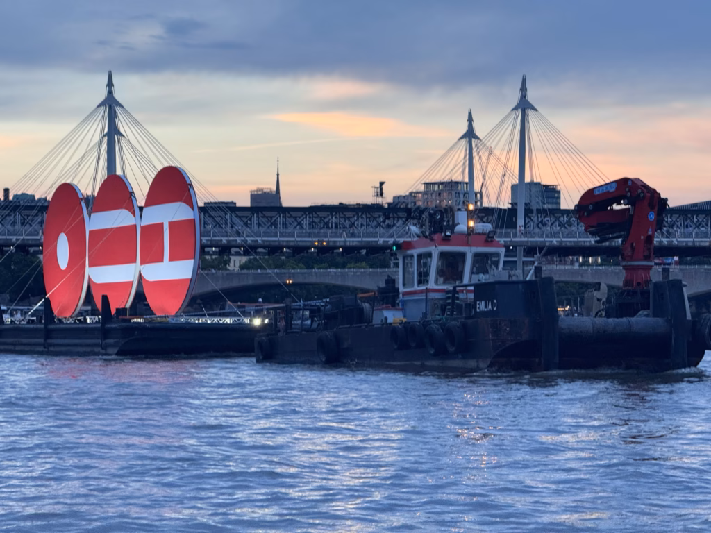 The BTS ARIRANG installation on the Thames, three lit red discs on a barge under tow past Hungerford Bridge and the Golden Jubilee Bridges at sunset