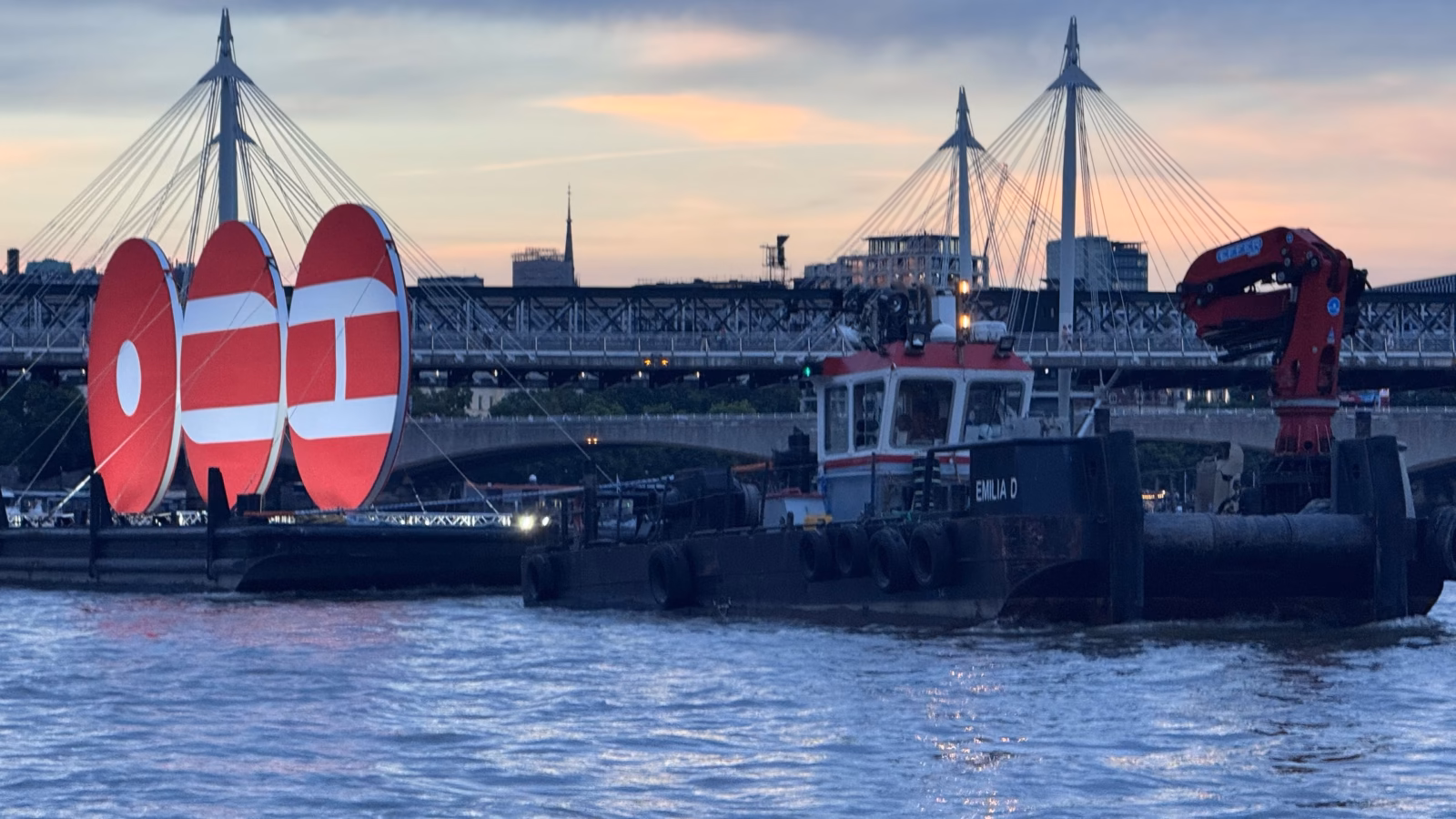 The BTS ARIRANG installation on the Thames, three lit red discs on a barge under tow past Hungerford Bridge and the Golden Jubilee Bridges at sunset