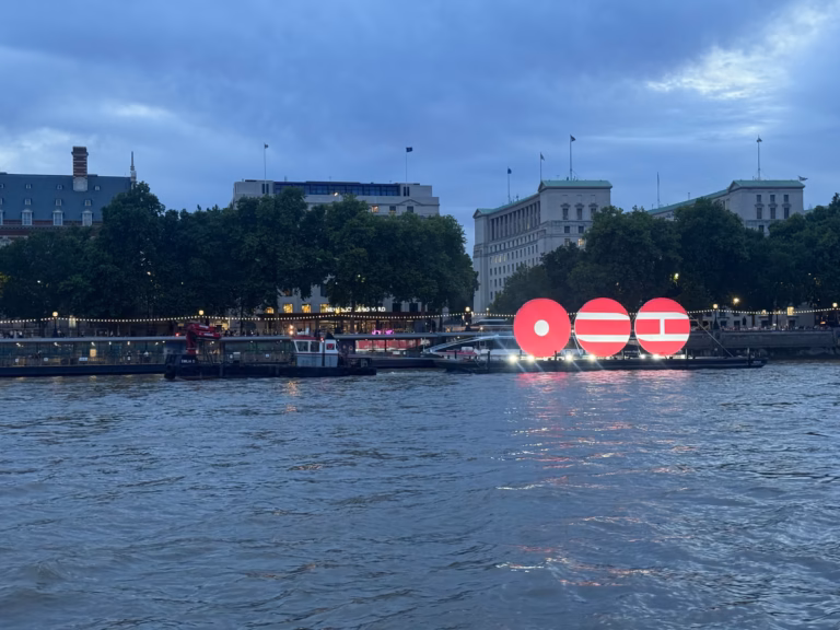 The BTS ARIRANG installation lit up on the Thames off Victoria Embankment at dusk, crowds along the riverside