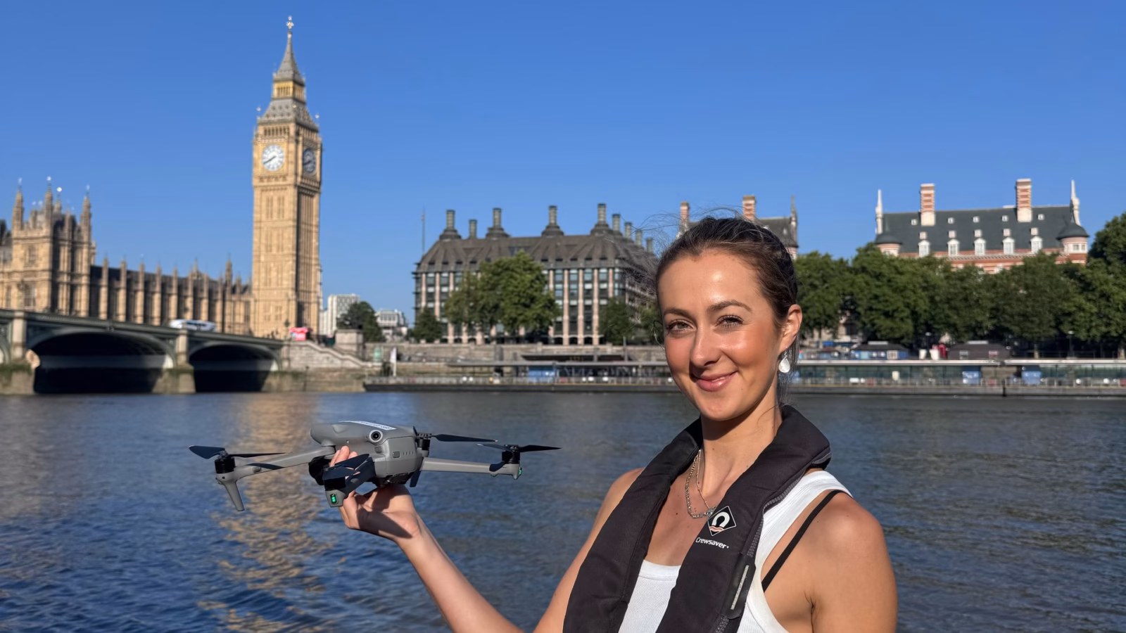 Drone operator Ella Goldsmith holding her drone and controller on our RIB on the Thames, with Big Ben, the Houses of Parliament and Westminster Bridge behind