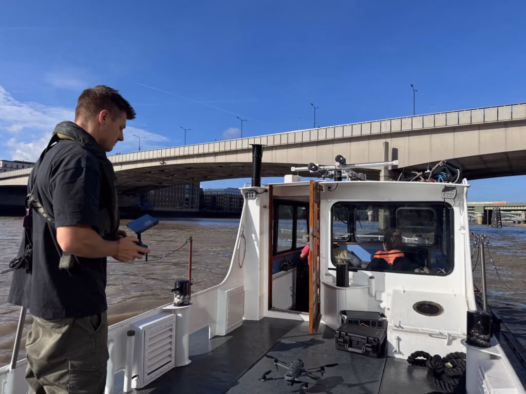 Drone pilot with a controller on the aft deck of a boat on the Thames, London Bridge behind and a drone on the deck