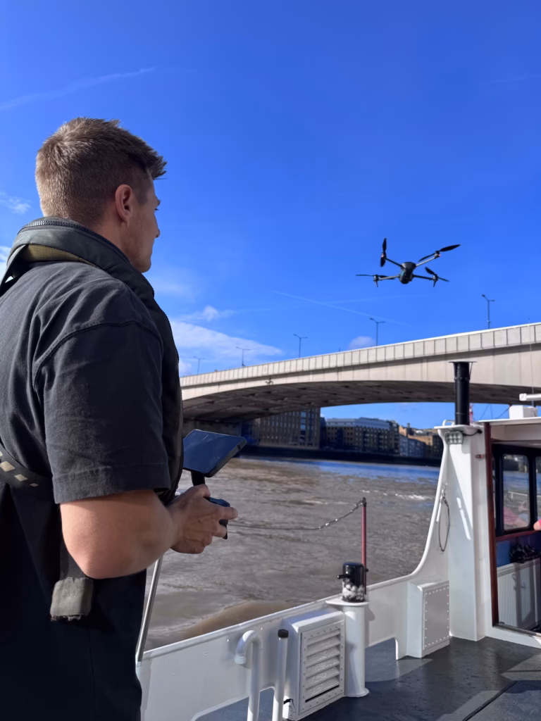 Drone pilot on the aft deck of a boat on the Thames, flying a drone overhead with London Bridge behind