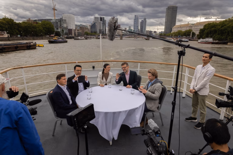 Roundtable set on the open deck of a passenger boat on the Thames, cameras rigged and the London skyline behind